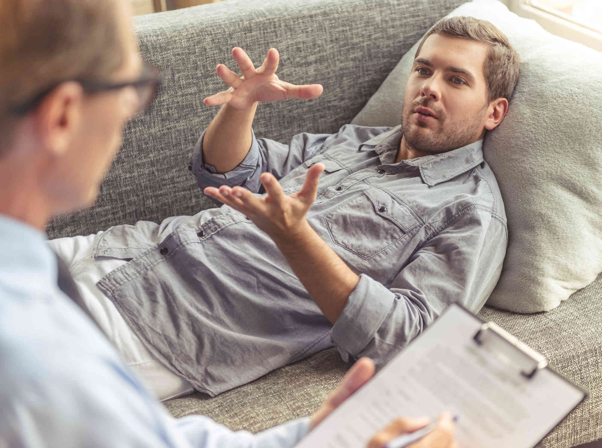 A man in a grey button down shirt lays on his back on a couch and talks to the therapist sitting next to him during a therapy session.